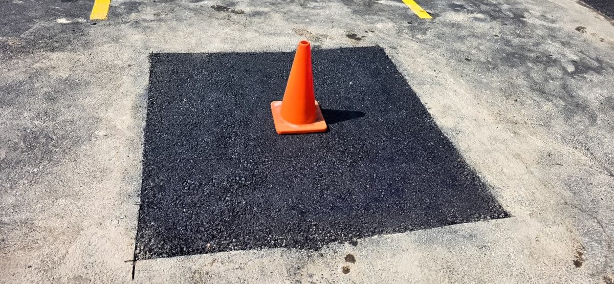 Rectangular black asphalt patch set into a light concrete surface with an orange cone on top.