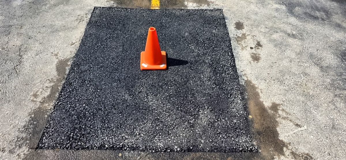 Compacted asphalt patch in a concrete pad marked with a single orange traffic cone.
