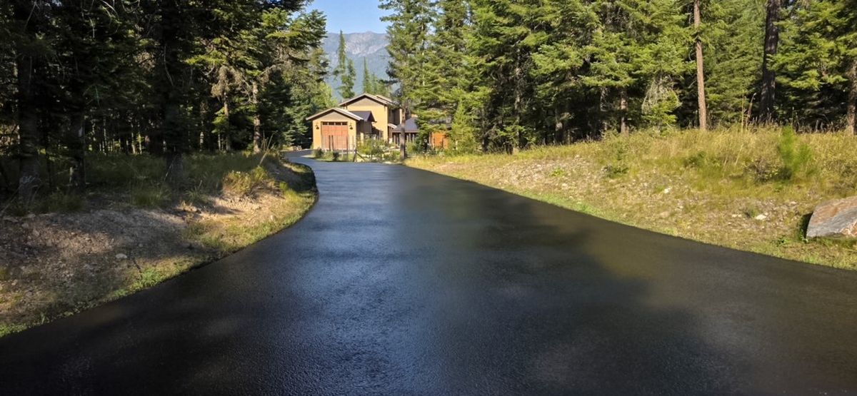 Freshly coated asphalt driveway curving through pine trees toward a house.
