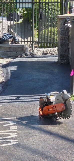 New asphalt at a gated driveway entrance tying into an existing paved street surface, with a walk-behind saw in the foreground.