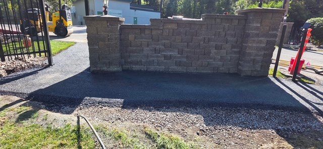 Fresh asphalt apron laid up to a stone block wall at a gated entrance, bordered by a gravel shoulder.