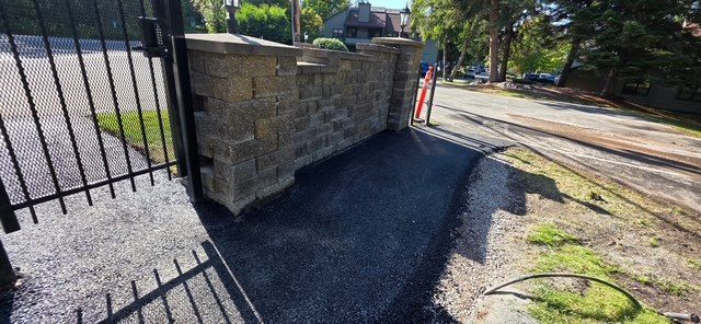Paved asphalt entrance section beside a stone wall, with the pavement edge meeting a gravel shoulder and open gate.