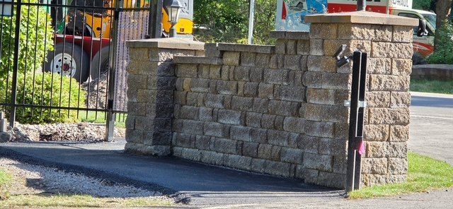 Finished black asphalt surface along a stone wall and gate post at a property entrance.