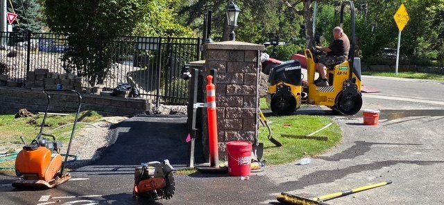 Active paving work area at a gated entrance with a ride-on roller, plate compactor, cones and hand tools in use.
