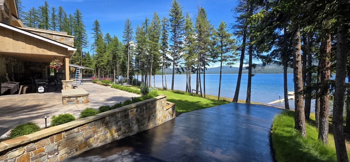 Freshly coated driveway running past a stone wall toward a lake framed by pine trees.