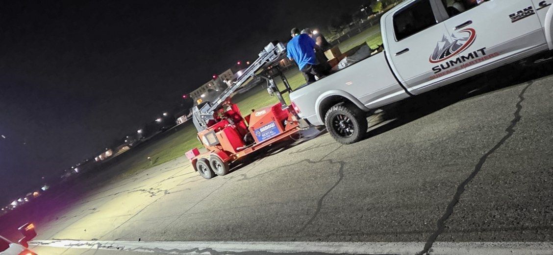 White service truck towing a red striping rig on a parking lot at night.