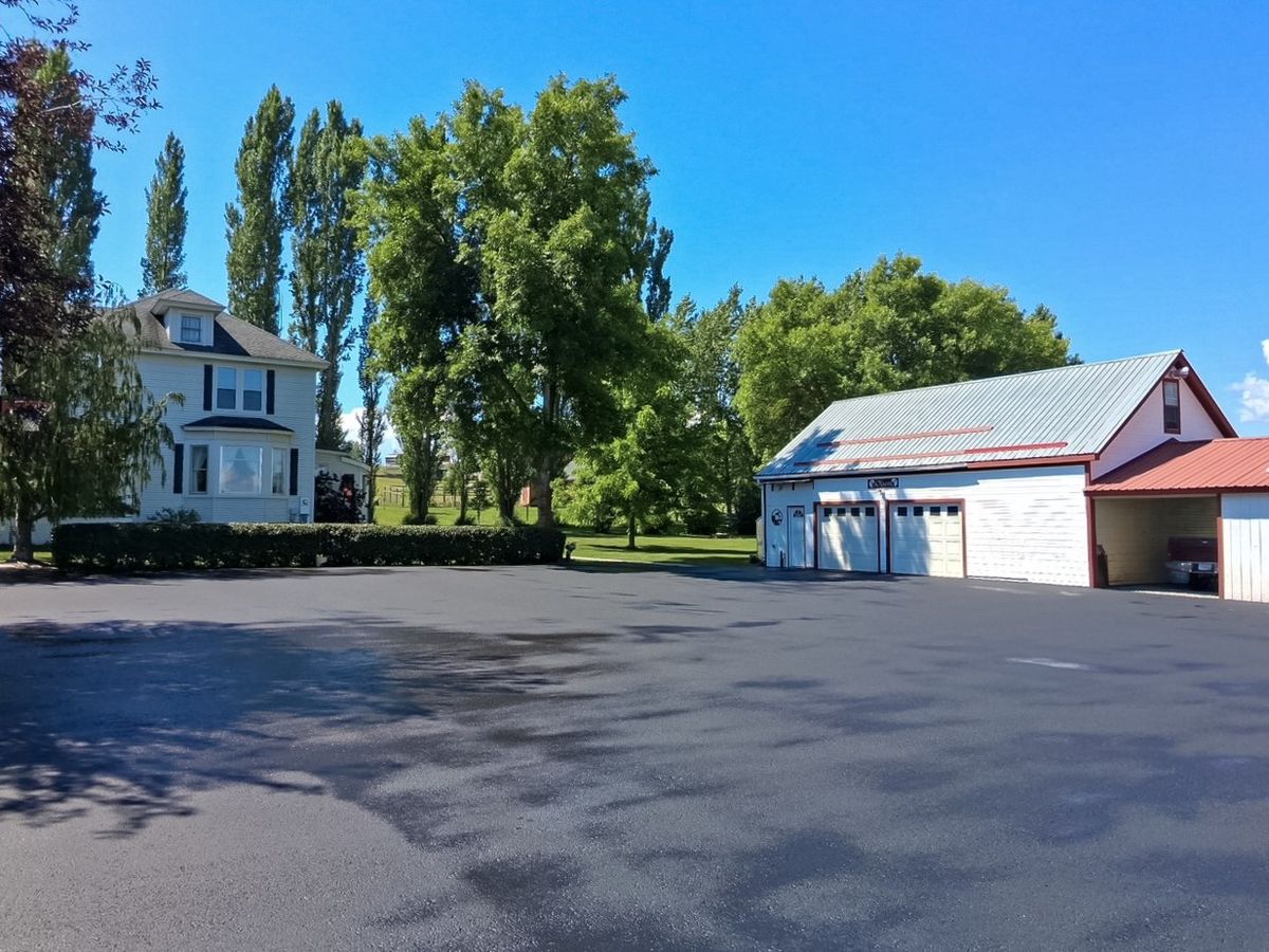 Freshly coated asphalt yard surface in front of a white house and red-roofed metal outbuildings.