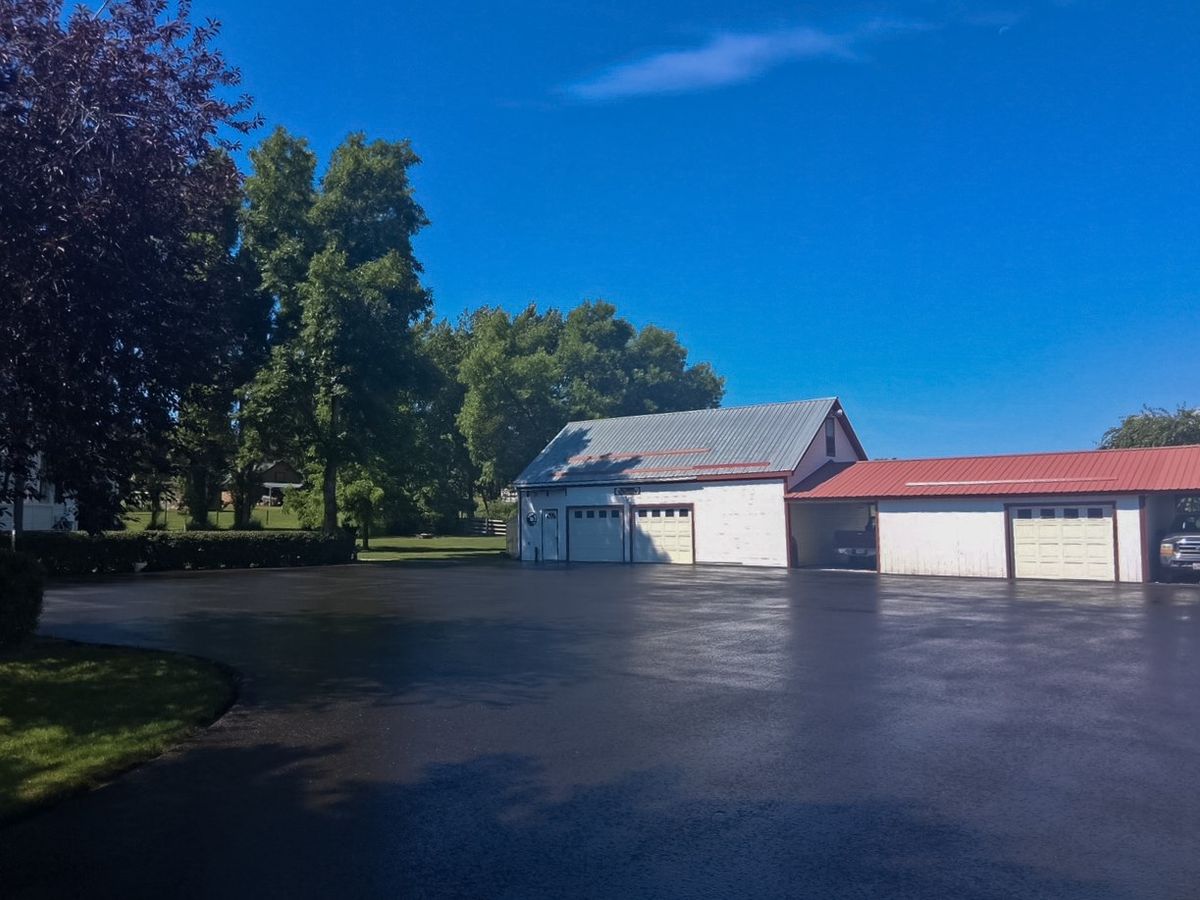 Freshly coated asphalt apron running along a row of white garage bays on a rural property.