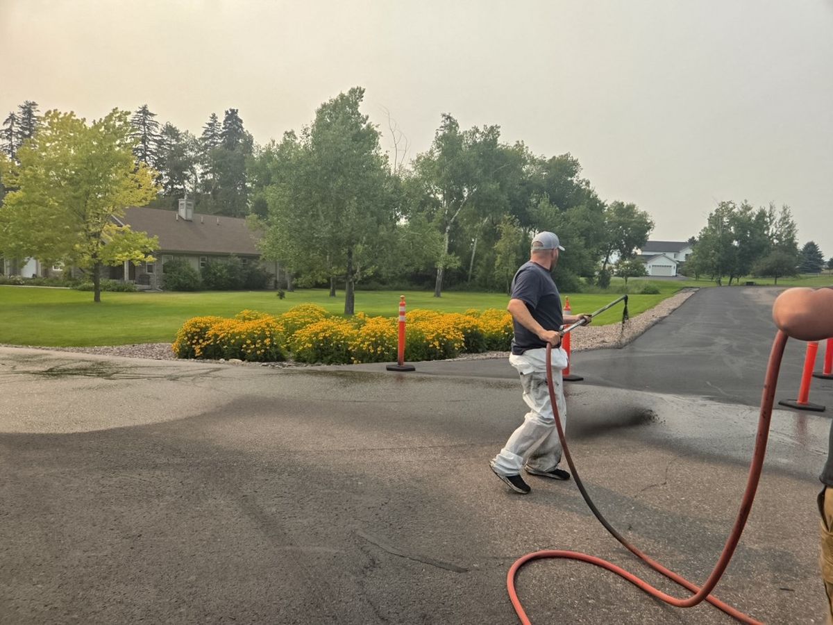 Worker applying sealer along a driveway approach marked off with orange traffic cones.