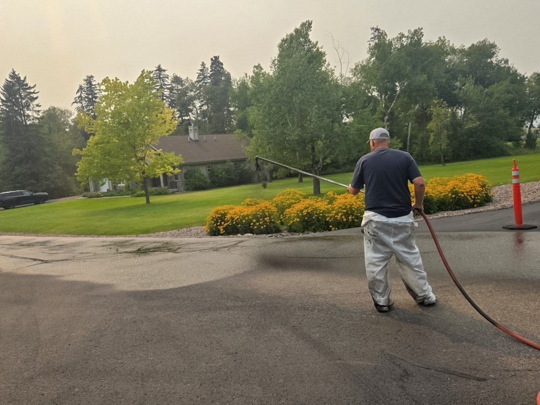 Worker spraying sealer along a residential driveway approach next to a flowering landscaped bed.