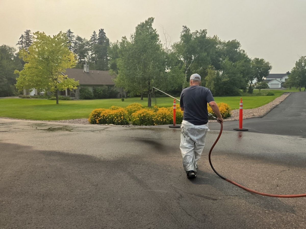 Worker moving a sealer wand along the shoulder of a paved residential approach with cones in place.