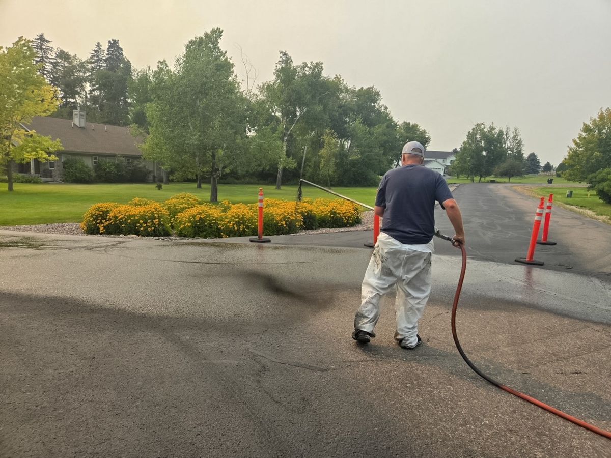 Coned-off lane beside a worker applying sealer on a residential paved approach.