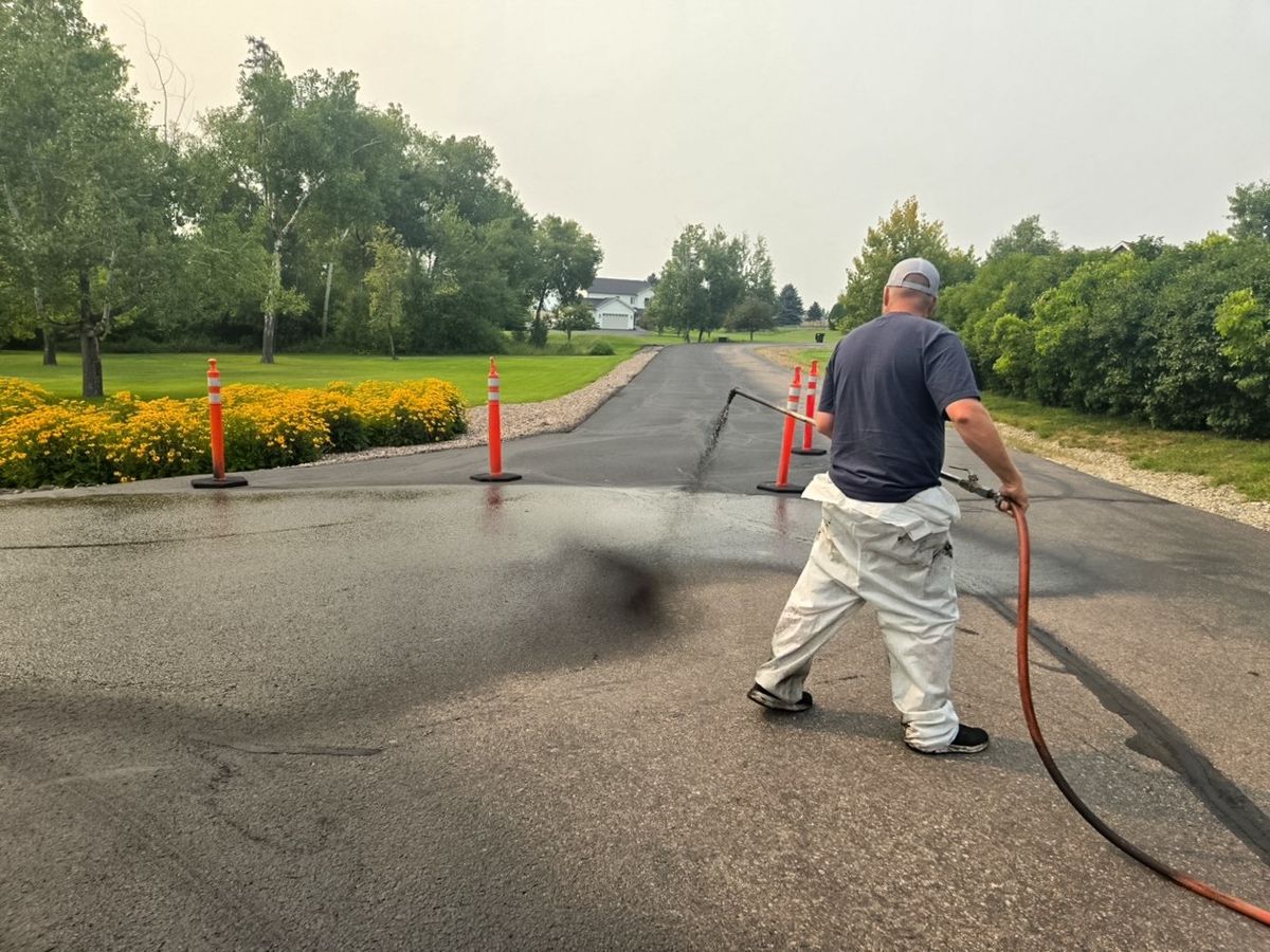 Worker applying sealer across the full width of a residential paved approach lined with cones.