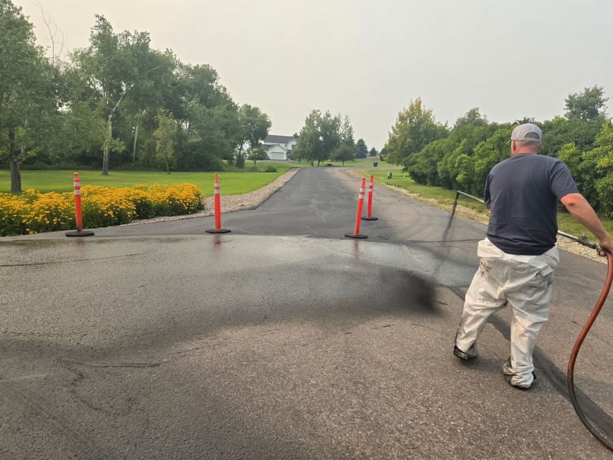 Worker making a finishing sealer pass beside orange cones on a residential paved approach.