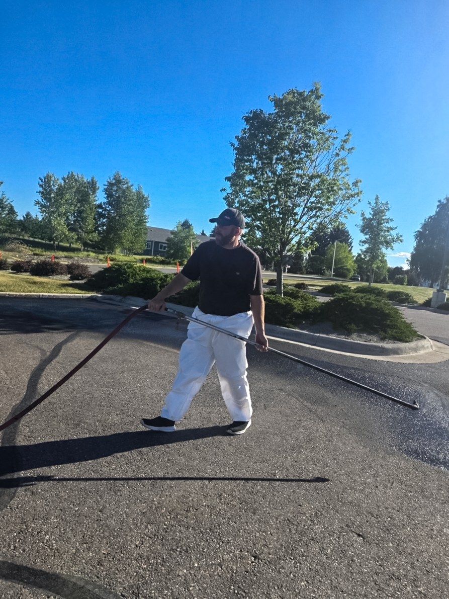 Worker applying sealer with a hand wand and hose across an asphalt driveway surface.