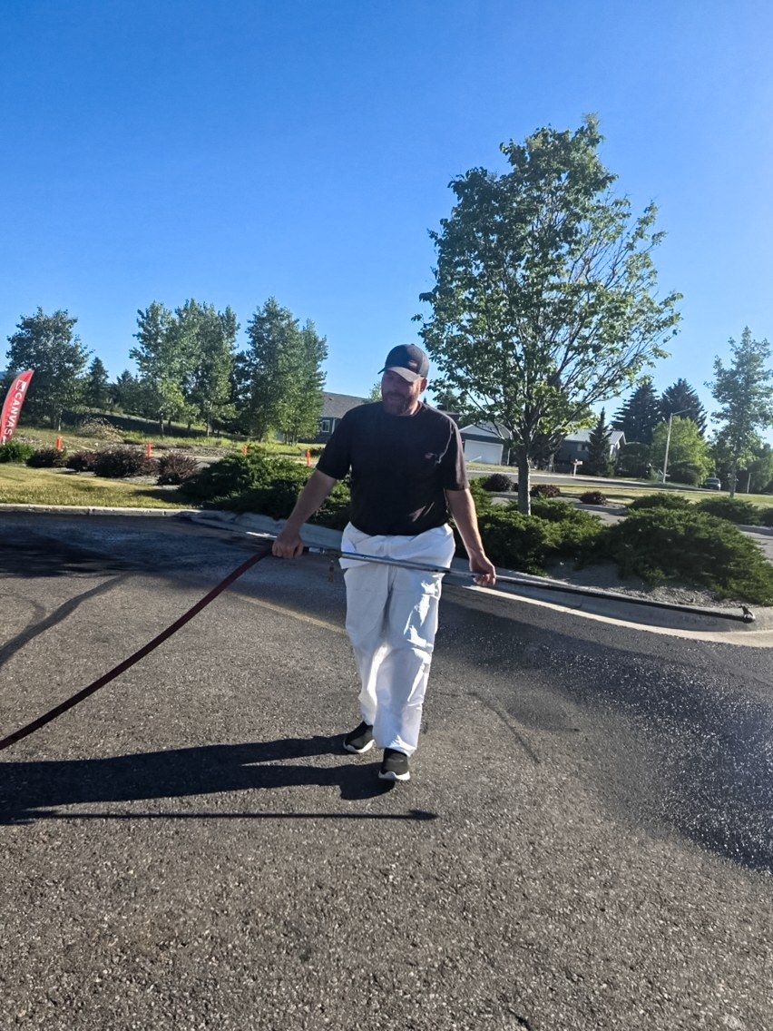 Worker walking a sealer wand along the edge of an asphalt driveway with the hose trailing behind.