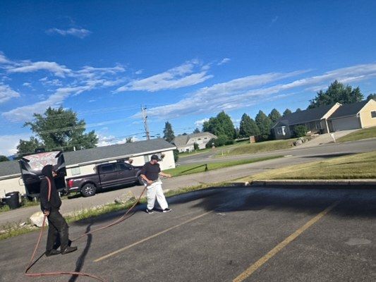 Worker spraying black sealer from a hose wand along a residential street, with a second crew member alongside.