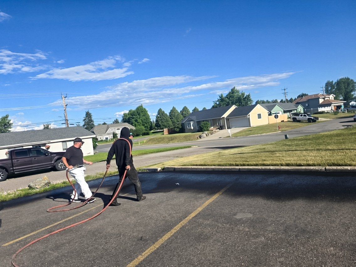 Two workers moving a sealer hose across a wide residential street with houses in the background.