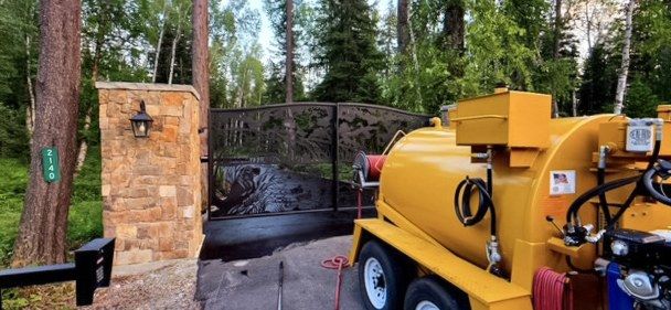 Yellow tow-behind sealer unit parked at a gated driveway entrance beside a stone pillar.
