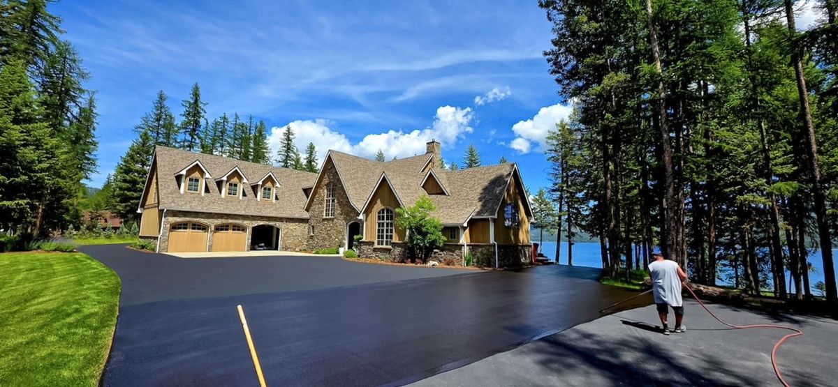 Worker applying sealer on a long residential driveway running downhill through tall pine trees.